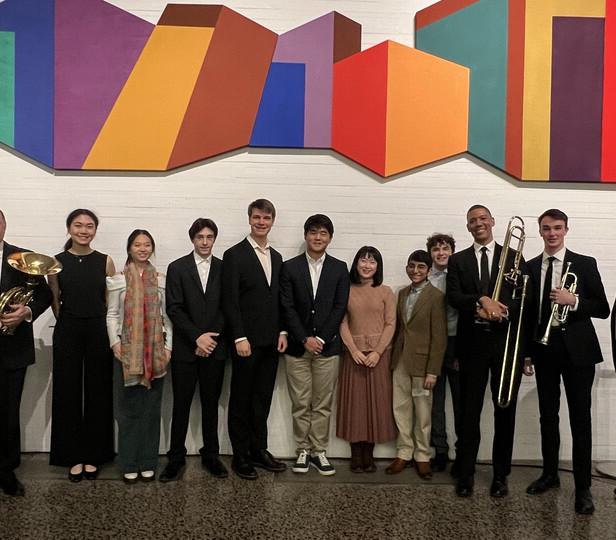 A group of people, including musicians holding instruments, stands in front of a colorful geometric wall art, smiling for a photo